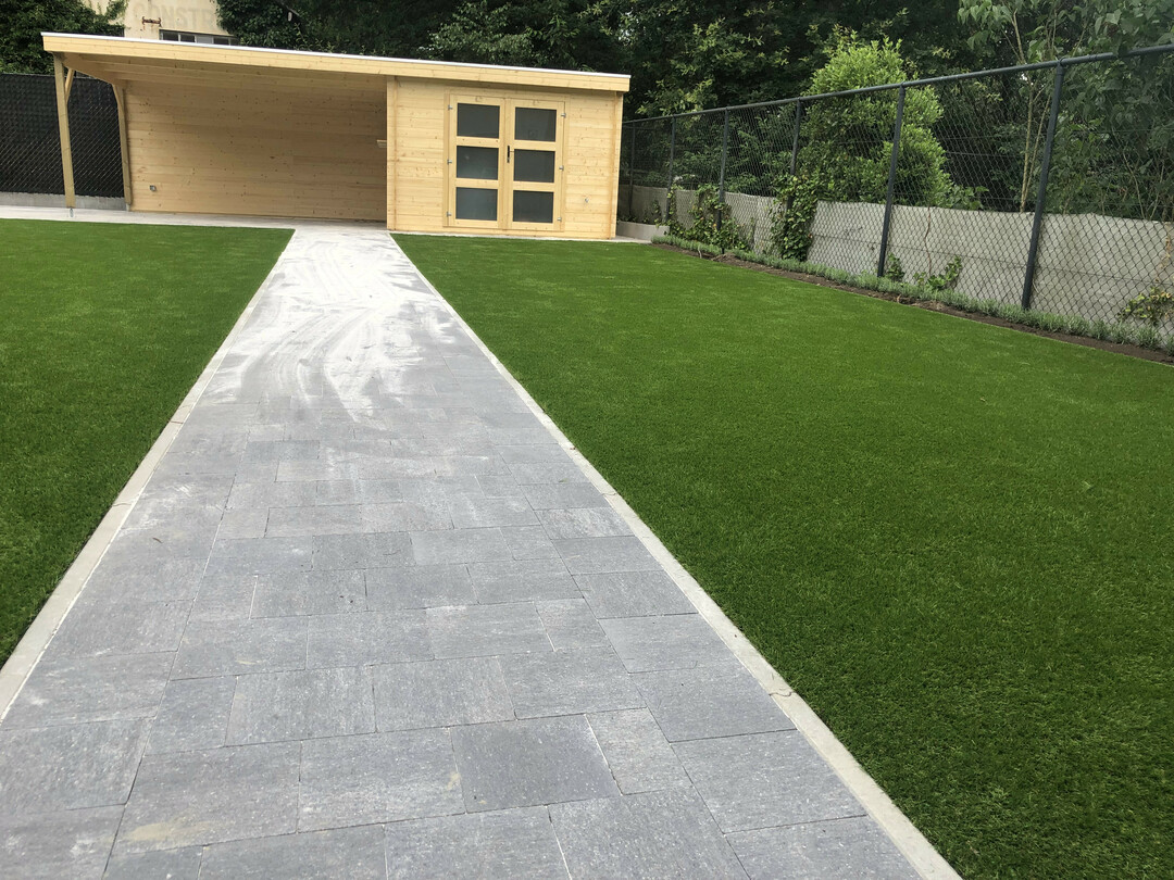breed tuinhuis met een lounge gedeelte, dit achter een klinkerpad omringd door kunstgras